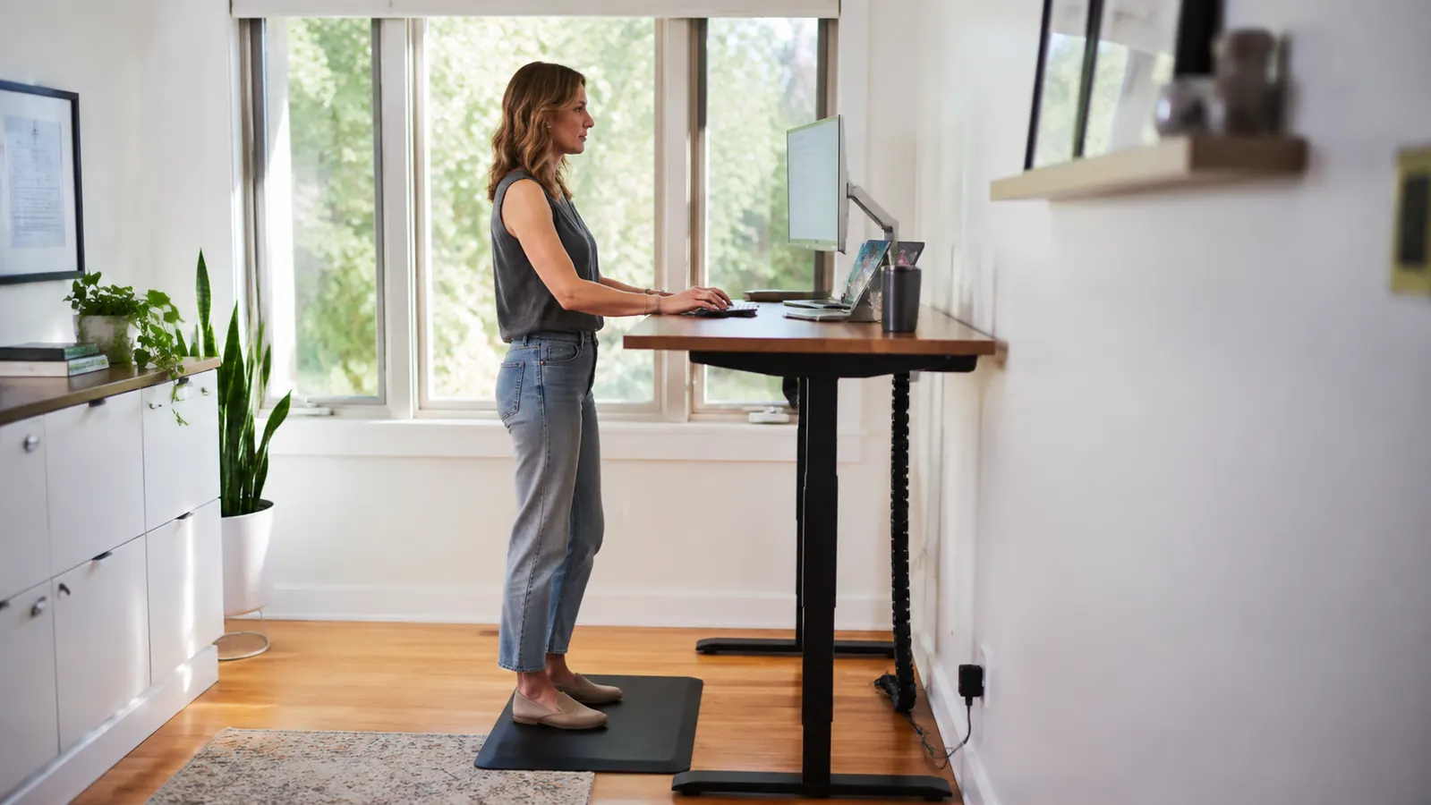 Person standing at a wood-top standing desk in a bright home office with a black anti-fatigue mat underfoot